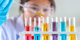 A woman is filling a liquid into a test tube using a pipette. Several test tubes with colorful liquids are in front of her.